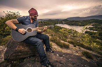 Outdoorsy musician with sunglasses and beanie plays guitar by a tranquil lake. HD wallpaper