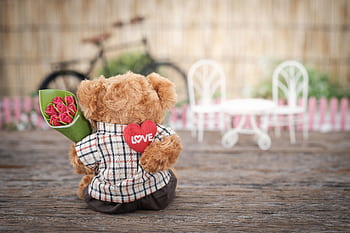 A cute teddy bear holding a heart with 'LOVE' and roses, set outdoors with rustic charm. HD wallpaper