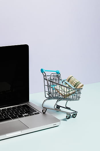 Mini shopping cart with rolled cash next to a laptop, representing online shopping. HD phone wallpaper