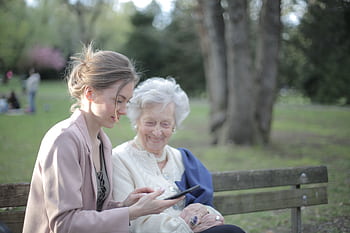 A young caregiver helps an elderly woman use a tablet in a peaceful park setting. HD wallpaper