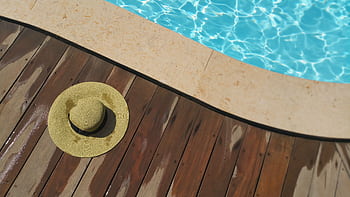 A straw hat rests on a wooden deck beside a sparkling blue swimming pool, perfect for summer relaxation. HD wallpaper