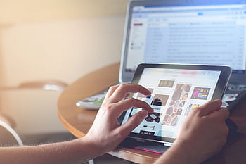 Hands using a tablet beside a laptop in a home office setting, showcasing online browsing. HD wallpaper
