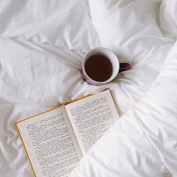 An inviting setup with a warm cup of tea and open book on white bedding, ideal for relaxation. HD wallpaper