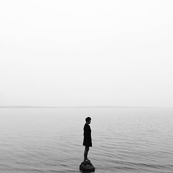 Black and white of a solitary person on a rock in a calm lake, evoking solitude and reflection. HD phone wallpaper