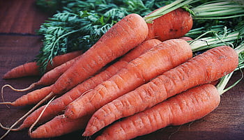 Close-up of fresh organic carrots with greens on a wooden surface, showcasing vibrant orange color. HD wallpaper
