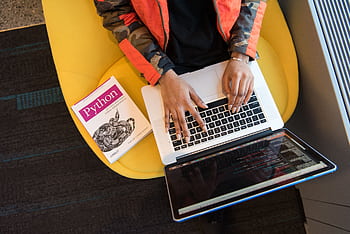 A developer typing code on a laptop with a Python book beside in an office. HD wallpaper