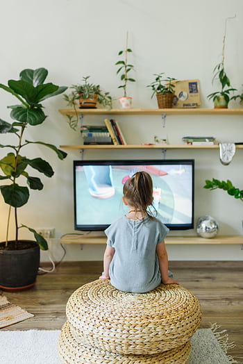 Child sitting on pouf in a cozy living room watching TV, surrounded by plants. HD phone wallpaper