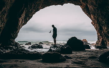 Silhouette of a person standing in a sea cave at Malibu beach, with ocean waves and rocks creating a dramatic scene. HD wallpaper