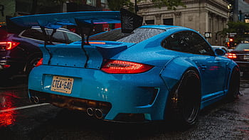 Dynamic blue sports car with large spoiler parked on a rainy city street, creating a dramatic urban ambiance. HD wallpaper