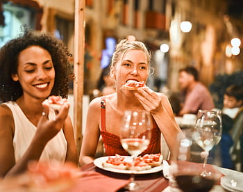 Two women enjoying bruschetta and wine at a vibrant outdoor restaurant. HD wallpaper