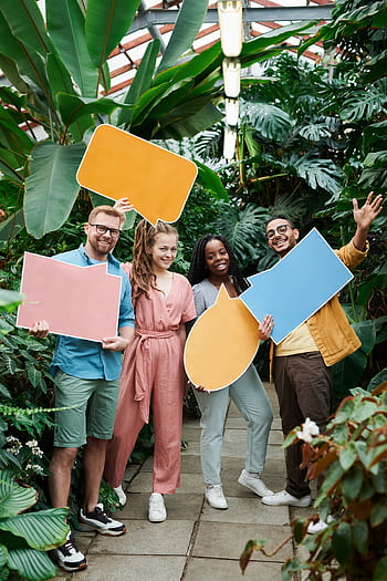 Cheerful group of friends holding colorful speech bubbles in a lush greenhouse setting. HD phone wallpaper