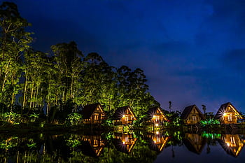 Stunning lakeside huts reflect in water under a night sky surrounded by tall trees. HD wallpaper