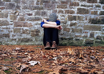 A solitary figure sits against a stone wall surrounded by fallen leaves, expressing isolation. HD wallpaper