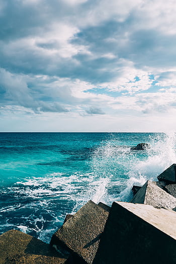 Powerful ocean waves crash against rocky barriers under a cloudy sky, showcasing nature's raw beauty. HD phone wallpaper