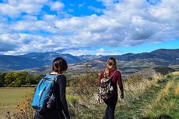 Two women hiking on a mountain trail with scenic views under a blue sky. HD wallpaper