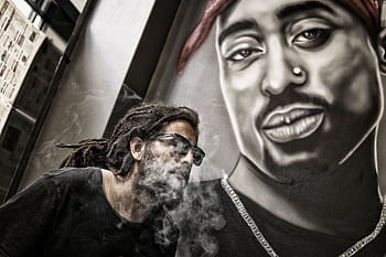 A man smoking in front of a mural featuring a hip hop artist, blending urban style and art. HD wallpaper