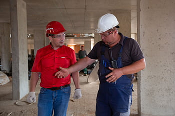 Two construction workers in safety gear discussing plans at a construction site. HD wallpaper