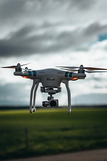 A drone flying over a green field under a dramatic cloudy sky, showcasing technology in nature. HD phone wallpaper