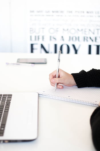 Close-up of a person's hand writing notes in an office setting with a laptop. HD phone wallpaper