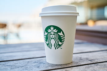 A disposable coffee cup with a lid outdoors on a wooden table, featuring a popular global logo. HD wallpaper