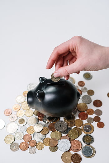 Hand inserting coin into black piggy bank among scattered coins, symbolizing savings and financial planning. HD phone wallpaper