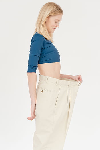 Young woman in oversized pants showing weight loss success, smiling in a studio. HD phone wallpaper