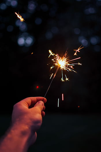 A person holding a bright sparkler outdoors in a festive night setting. HD phone wallpaper