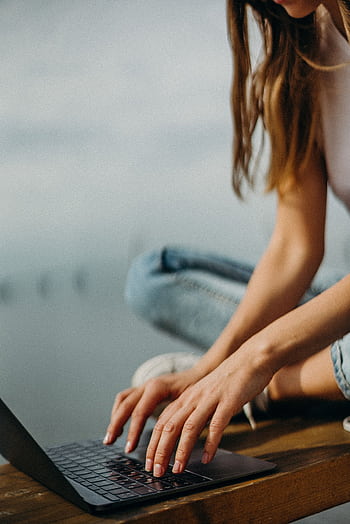 A young woman working remotely on her laptop by a calm waterfront. HD phone wallpaper