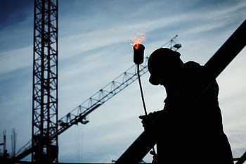 Silhouette of a construction worker using a blowtorch at a building site against a crane-filled skyline. HD wallpaper