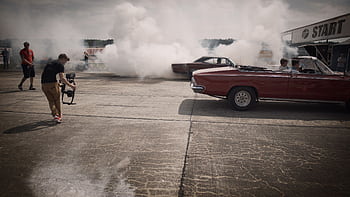 A dynamic scene of classic cars and filmmakers capturing a smoky drag race event. HD wallpaper