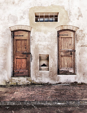 Two weathered wooden doors on an aged brick wall with a small barred window above, creating a rustic facade. HD phone wallpaper