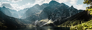 Serene mountain lake scene with rugged peaks and forested valleys under a bright day sky. HD wallpaper