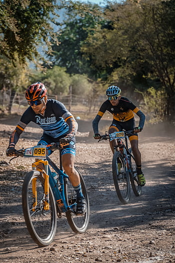 Two cyclists race fiercely on a dusty mountain trail under clear skies. HD phone wallpaper