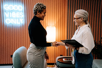 Two diverse businesswomen in a stylish office shaking hands under a 'Good Vibes Only' sign. HD wallpaper