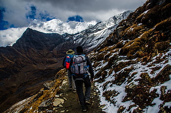 Two hikers navigating a snowy trail in rugged mountainous terrain under dramatic skies. HD wallpaper