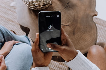 Close-up of hands holding a smart displaying the TikTok app interface on a wooden surface. HD wallpaper