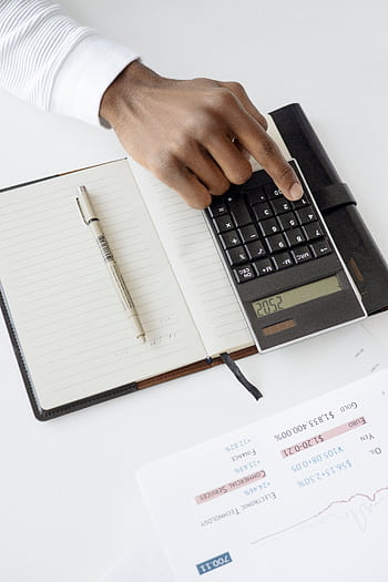 A hand using a calculator with financial documents and notebook, depicting an office setup for financial analysis. HD phone wallpaper