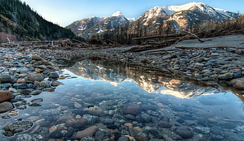 Tranquil winter scene with snow-capped mountains reflected in a serene stream, framed by stones and pebbles. HD wallpaper