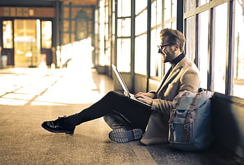 Man sitting by a window at a Budapest train station, working on his laptop with sunlight streaming through. HD wallpaper