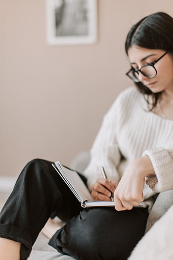 Woman sitting on a sofa, wearing glasses, writing in a journal, focused and relaxed. HD phone wallpaper