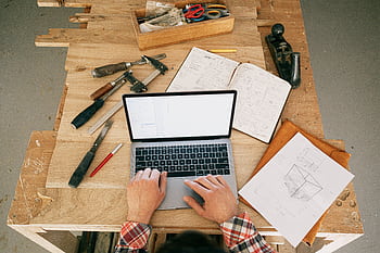 Carpenter using laptop for woodwork designs at a workbench in a workshop. HD wallpaper
