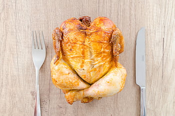 Top view of a perfectly roasted chicken placed between fork and knife on a wooden table. HD wallpaper
