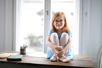 Smiling young girl with glasses sits cross-legged on a table indoors, looking cheerful. HD wallpaper