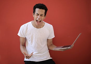 A man expressing frustration with his laptop against a red background, shouting and gesturing in studio. HD wallpaper