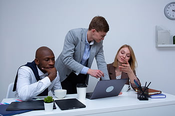 A diverse team collaborating in a modern office environment, discussing work on a laptop. HD wallpaper