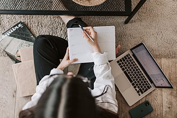 Overhead view of a woman writing in a planner beside a laptop in a cozy home setting. HD wallpaper