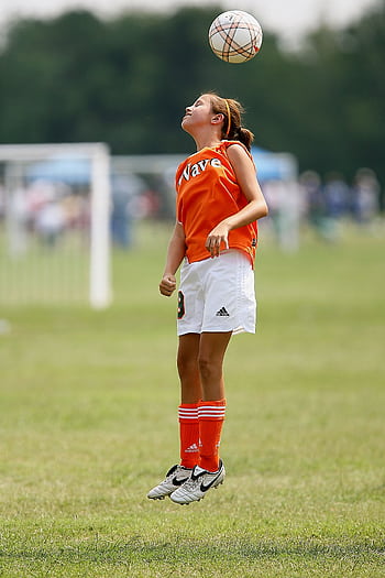 A young girl jumps to head a soccer ball during a game on a grassy field. HD phone wallpaper