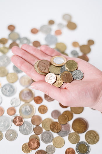 A hand holding a variety of coins with more scattered on a white surface, symbolizing currency and finance. HD phone wallpaper