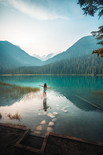 A woman stands peacefully in a clear Canadian mountain lake surrounded by lush greenery. HD phone wallpaper