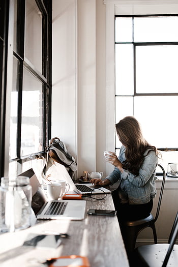 A young woman working on her laptop, sipping coffee in a bright café setting. HD phone wallpaper